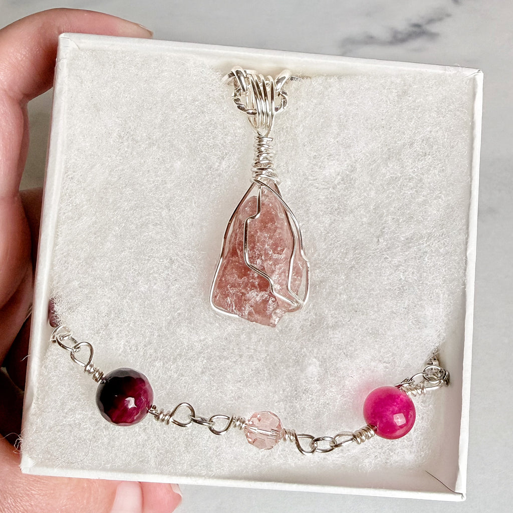 A pink crystal pendant necklace and matching beaded bracelet with silver wire wrapping, presented in a white gift box held by a hand.