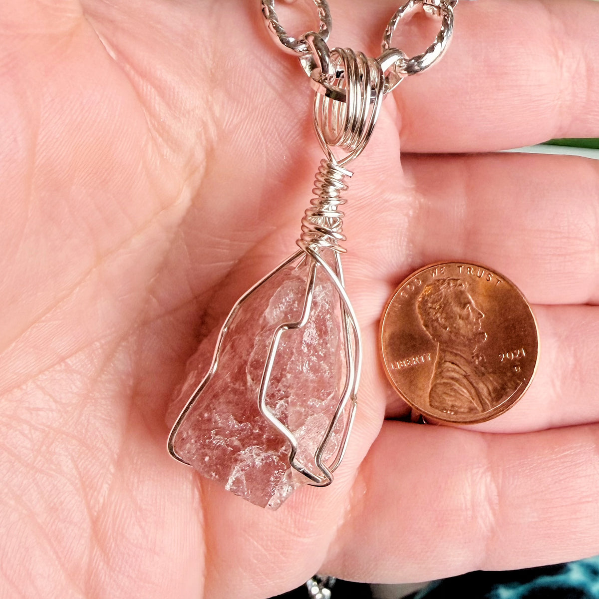 A close-up of a hand holding a raw pink stone pendant wrapped in silver wire, with a U.S. penny for scale.