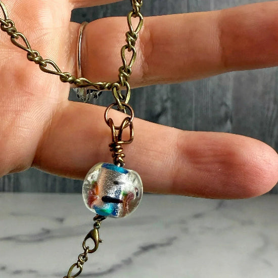 Necklace with a multicolored glass bead charm held by a hand on a marble surface.
