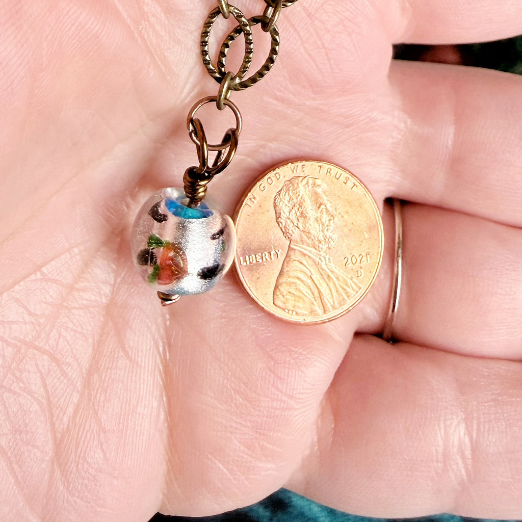 A close-up of a hand holding a small handmade glass bead charm on a bronze chain next to a US penny for scale.