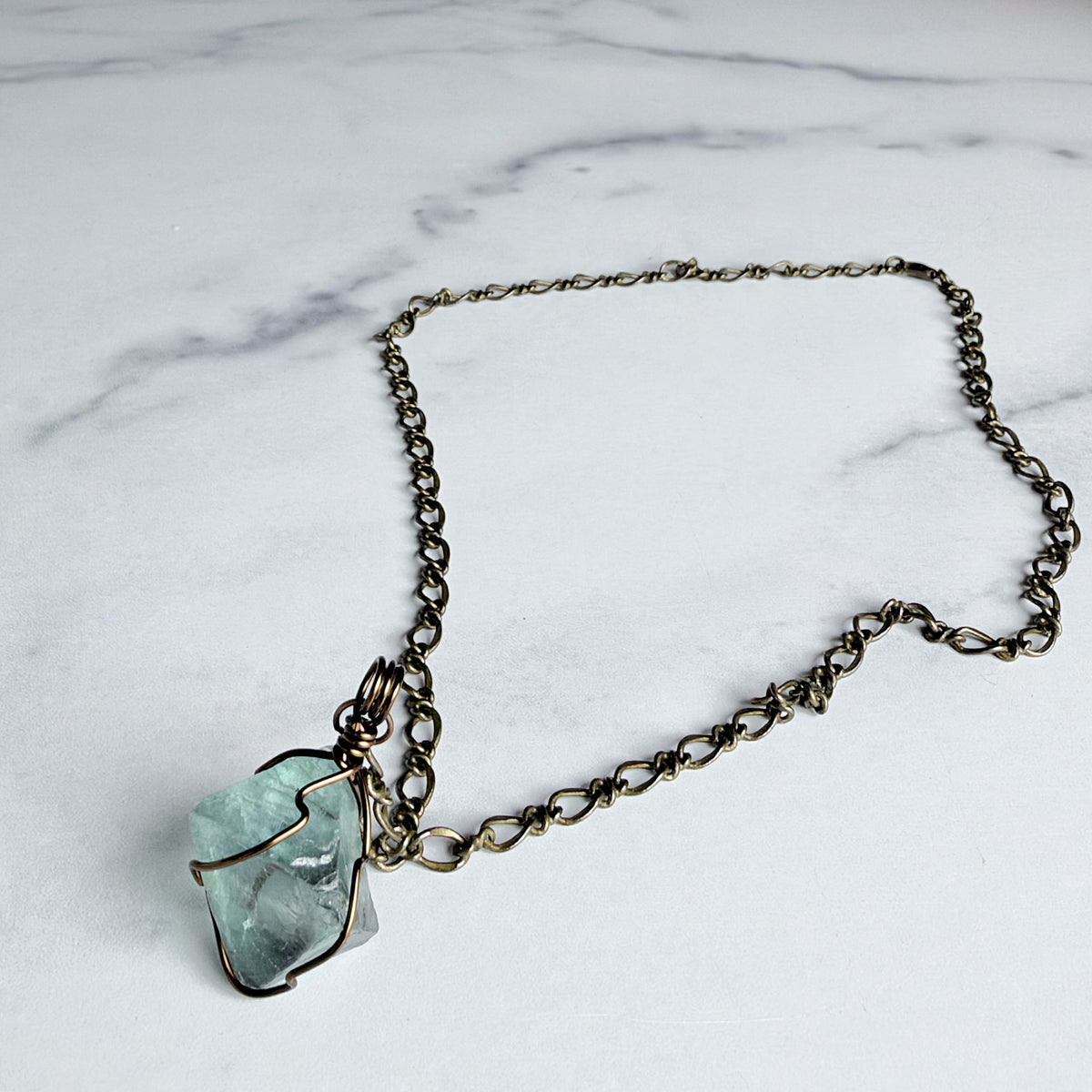 A handmade necklace featuring a raw, irregular light blue-green stone wrapped in dark bronze wire, suspended from a matching dark bronze chain, displayed on a white marble surface.