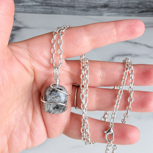 A hand holding a silver chain necklace with a round gray and white marbled stone pendant wrapped in silver wire, against a light marble background.