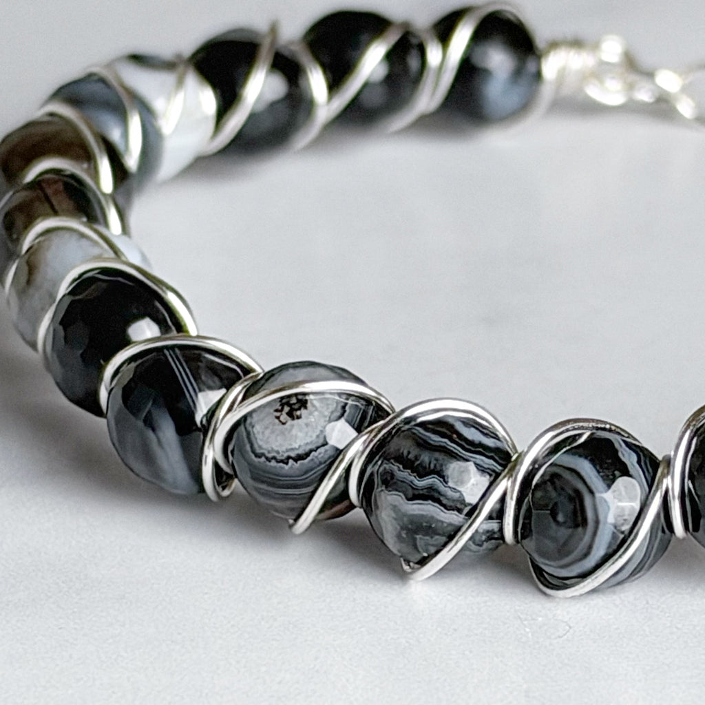 A close-up of a beaded bracelet featuring polished black and gray agate beads wrapped in silver-toned metal wire, on a light gray background.