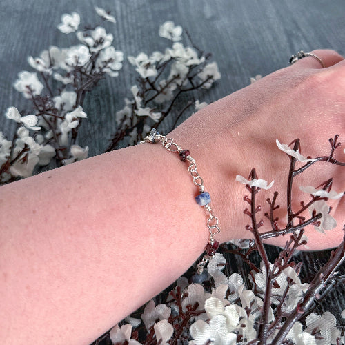 Hand wearing a bracelet with small stones, surrounded by white flowers on a dark background