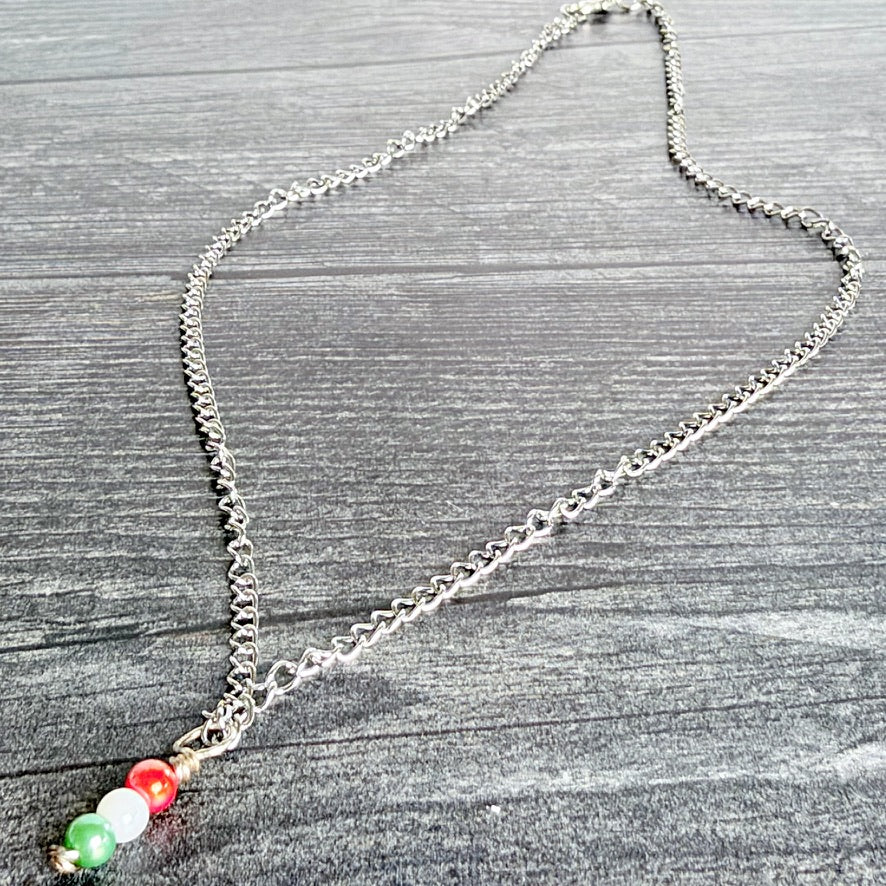A silver chain necklace with a pendant featuring three small beads in green, white, and red, arranged like the Italian flag, displayed on a gray wood-textured background.