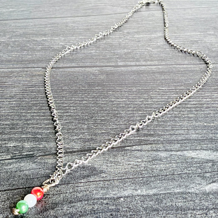 A silver chain necklace with a pendant featuring three small beads in green, white, and red, arranged like the Italian flag, displayed on a gray wood-textured background.