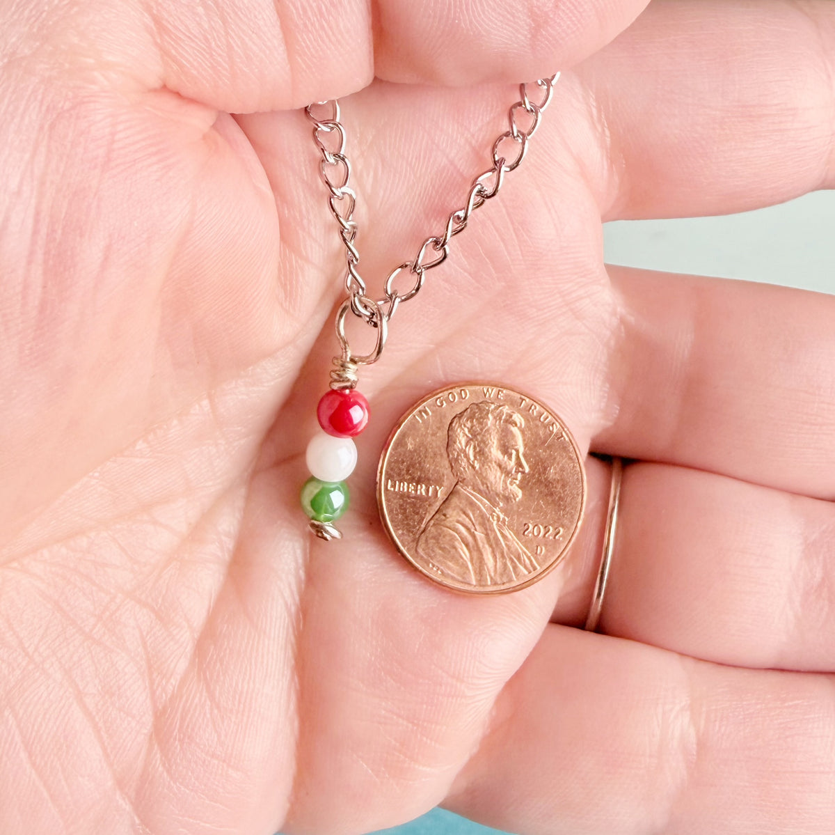 A close-up of a hand holding a silver chain necklace with a small charm made of red, white, and green beads, shown next to a U.S penny for scale