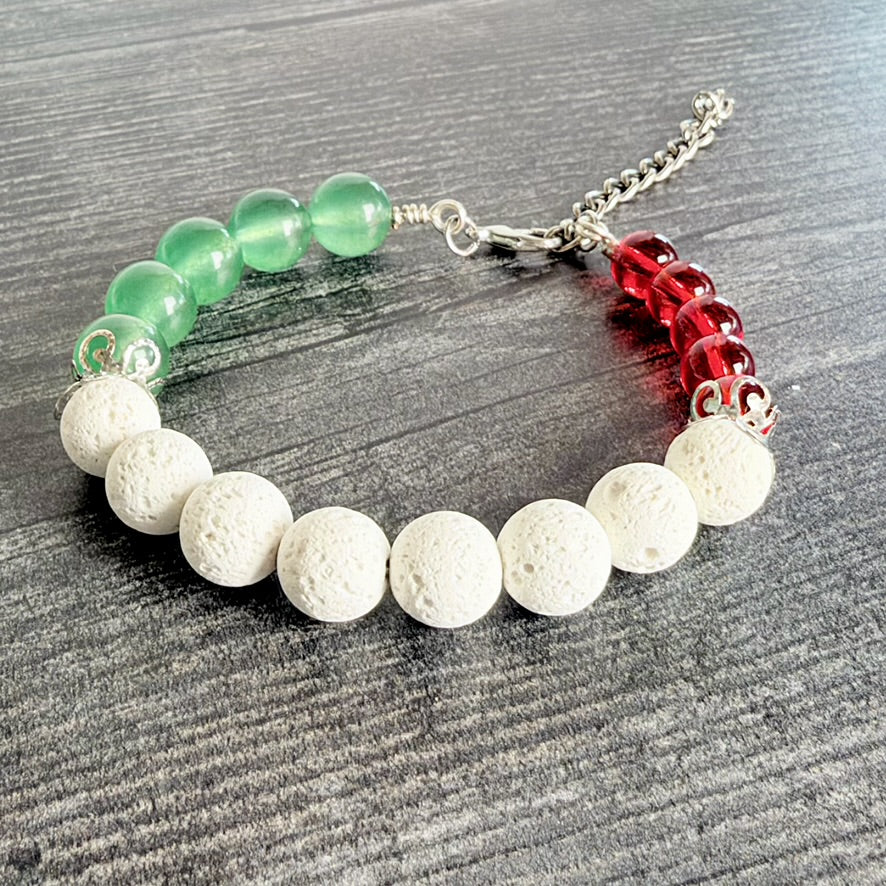 A beaded bracelet with white porous lava stone beads, green translucent round beads, and red translucent round beads, finished with a silver-tone chain extender, photographed on a dark wood surface.