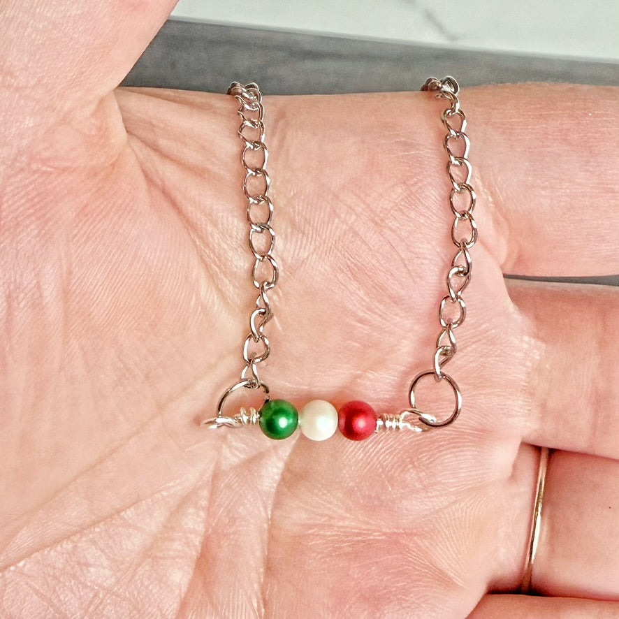 A silver chain bracelet with three small beads in green, white, and red, held on a person's palm.