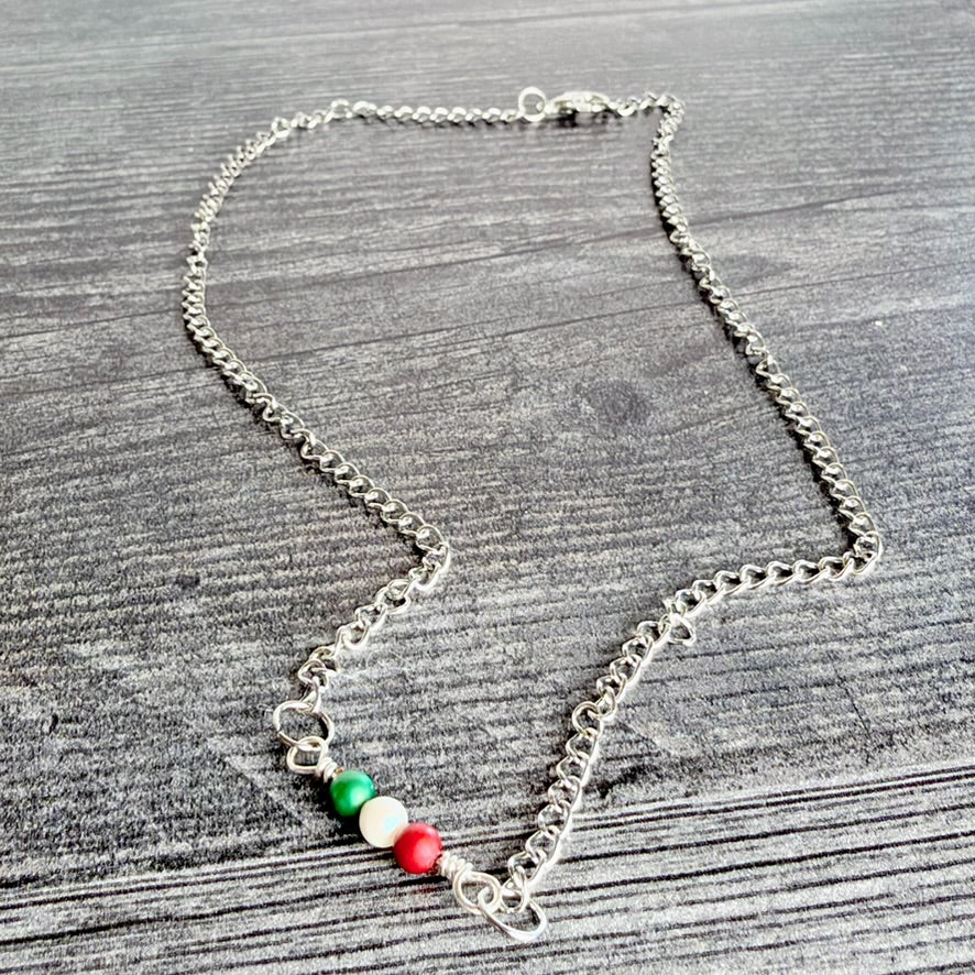 A silver chain necklace featuring three small beads in the colors of the Italian flag (green, white, and red) on a gray wood-textured background.