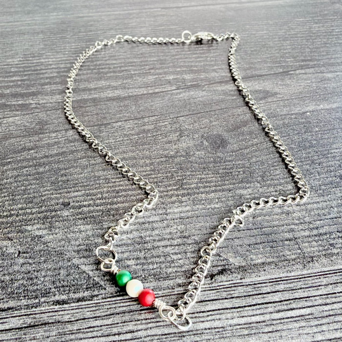 A silver chain necklace featuring three small beads in the colors of the Italian flag (green, white, and red) on a gray wood-textured background.