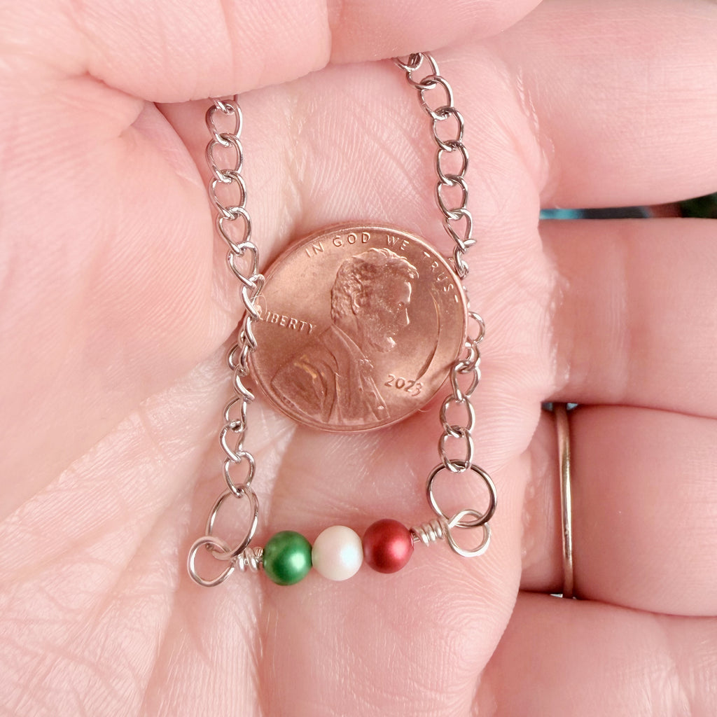 A close-up of a hand holding a silver chain necklace with a penny for scale and three small beads in green, white, and red.