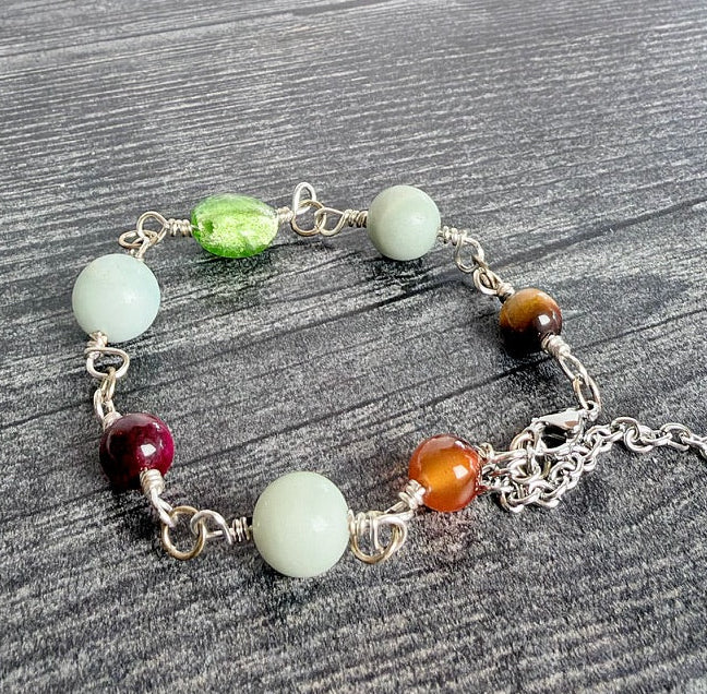 A beaded chakra bracelet with silver-toned metal links and extender chain, featuring round beads in light green, white, dark red, orange, and brown, displayed on a dark gray wood background.