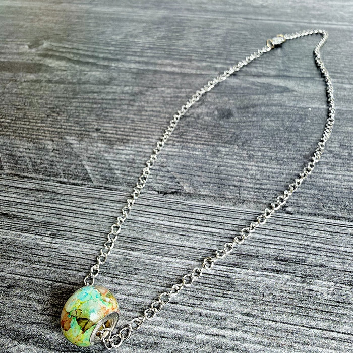 A silver chain necklace with a colorful, round, textured bead pendant featuring green, yellow, and orange tones, displayed on a gray wooden surface.