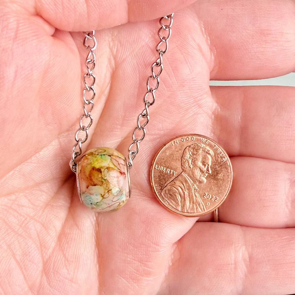 A close-up of a hand holding a silver chain necklace with a marbled, earthy-colored oval pendant next to a U.S.