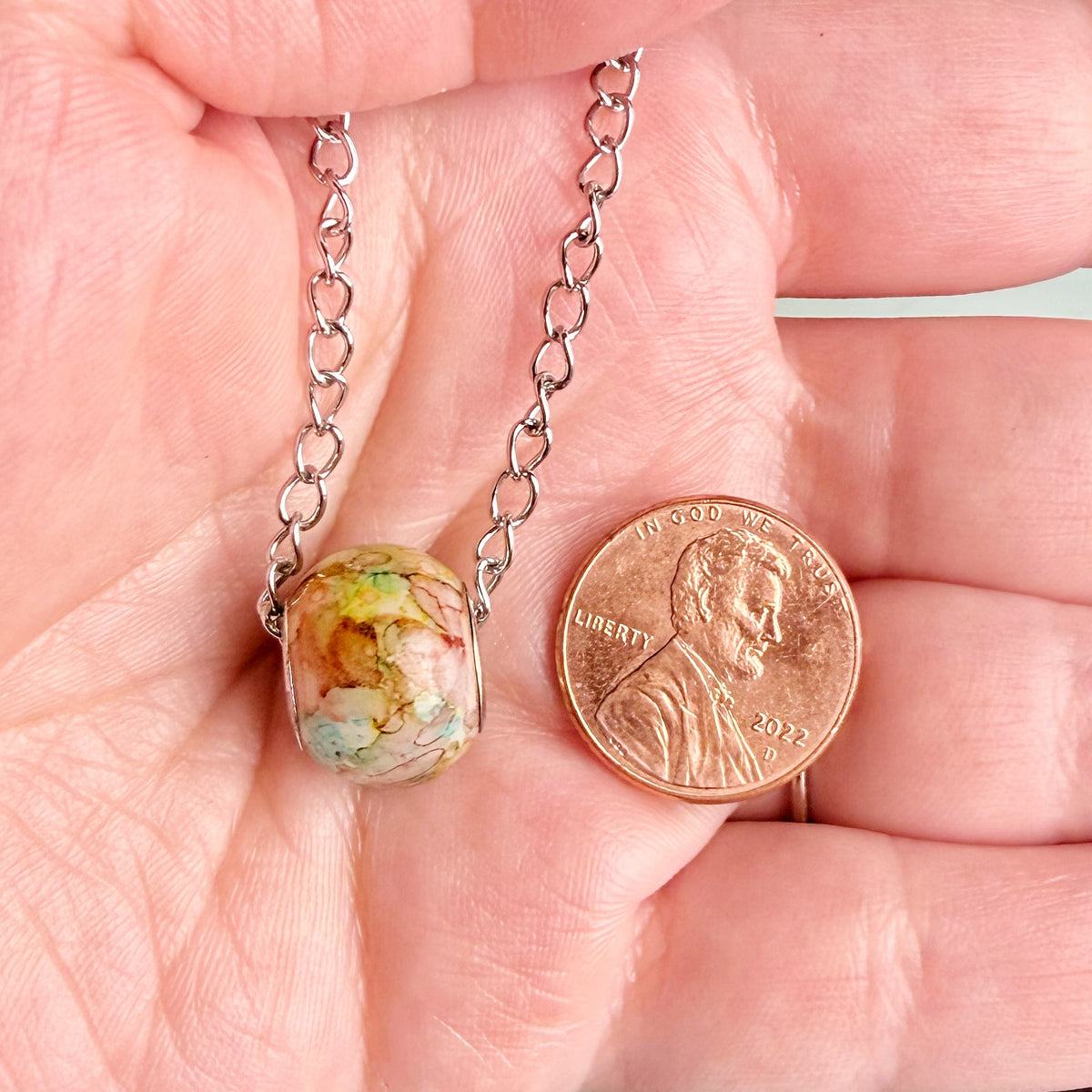 A close-up of a hand holding a silver chain necklace with a marbled, earthy-colored oval pendant next to a U.S.