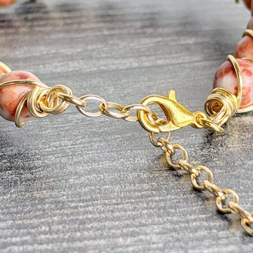 Close-up of a gold chain bracelet with a lobster clasp and extension chain, featuring pink and white marbled gemstone beads on a textured gray background.