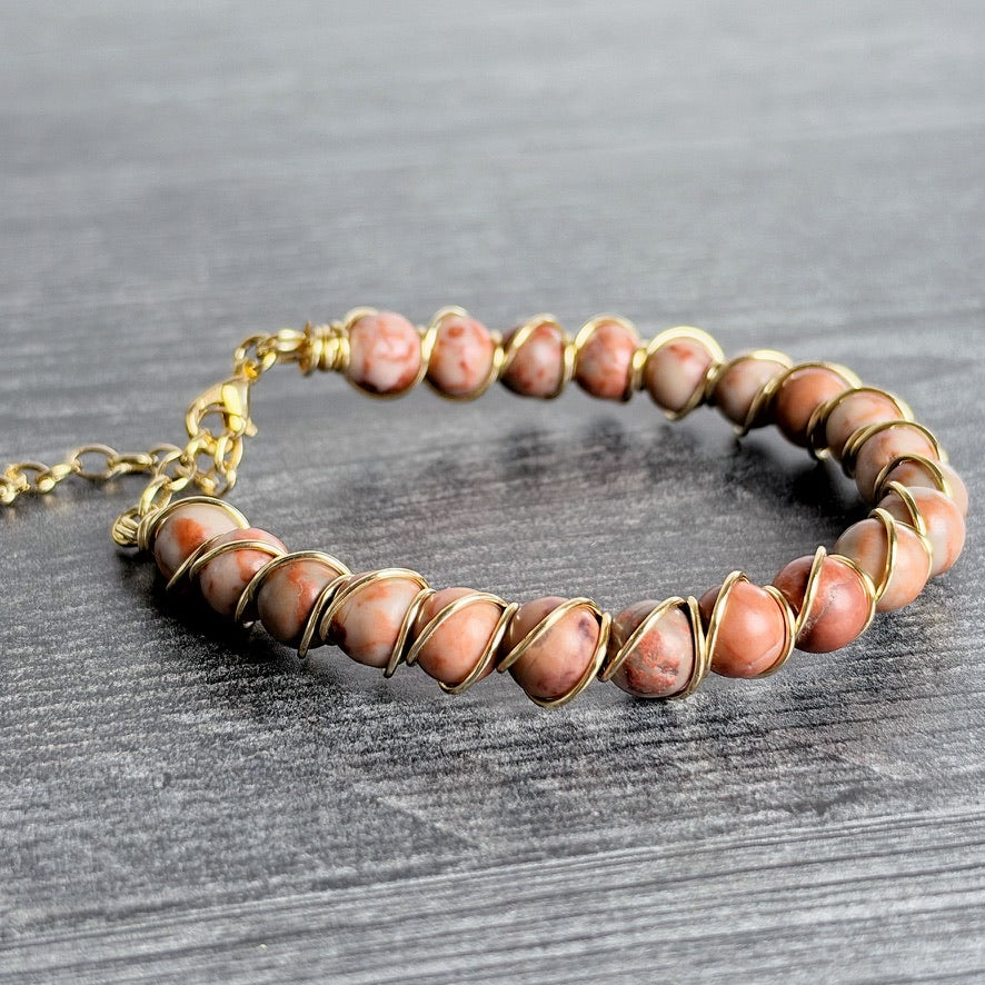 A handmade beaded bracelet featuring round pinkish-brown stone beads wrapped in gold-toned wire, displayed on a dark gray textured wooden surface.