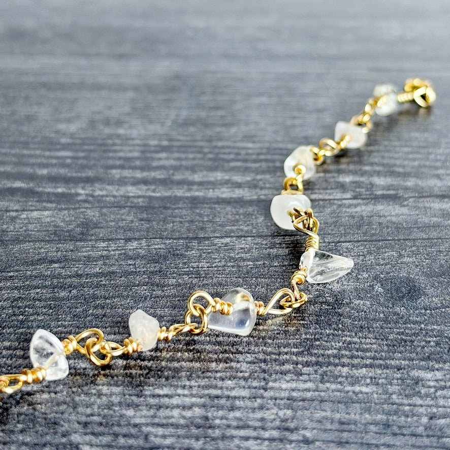 A gold chain bracelet with irregular, clear quartz crystal beads, displayed diagonally on a dark gray wood-textured background.