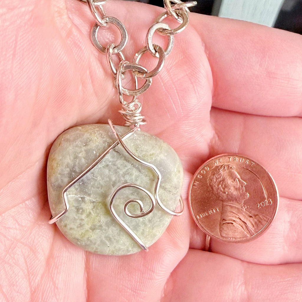 A handmade silver wire-wrapped stone pendant necklace held in a person's hand, shown next to a U.S. penny for scale.