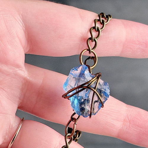 A close-up of a hand holding a bracelet with a blue faceted gemstone wrapped in bronze-toned metal, suspended from a matching chain.