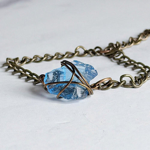 A handmade necklace featuring a raw, faceted light blue gemstone pendant wrapped in bronze-colored wire, suspended from a thick antique bronze chain on a white background.