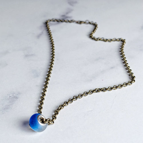 An antique bronze chain necklace with a small, round, blue glass bead pendant, displayed on a light gray marble surface.