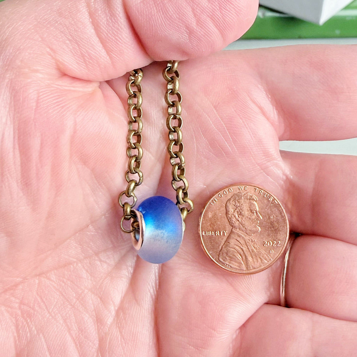 A close-up of a hand holding a small necklace with a blue glass bead and an antique bronze chain, shown next to a penny for scale.