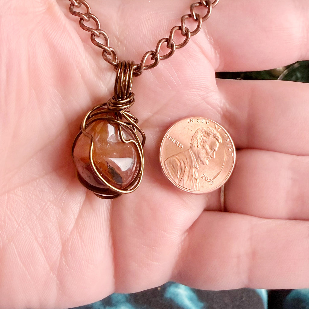 A handmade copper wire-wrapped pendant necklace with a faceted amber-brown stone, held in a person's hand next to a US penny for scale.