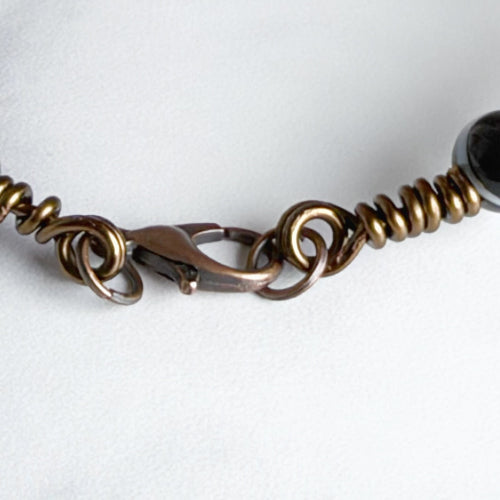 Close-up of a metal chain bracelet featuring a lobster clasp, coiled spring links, and a dark round bead on a white background.