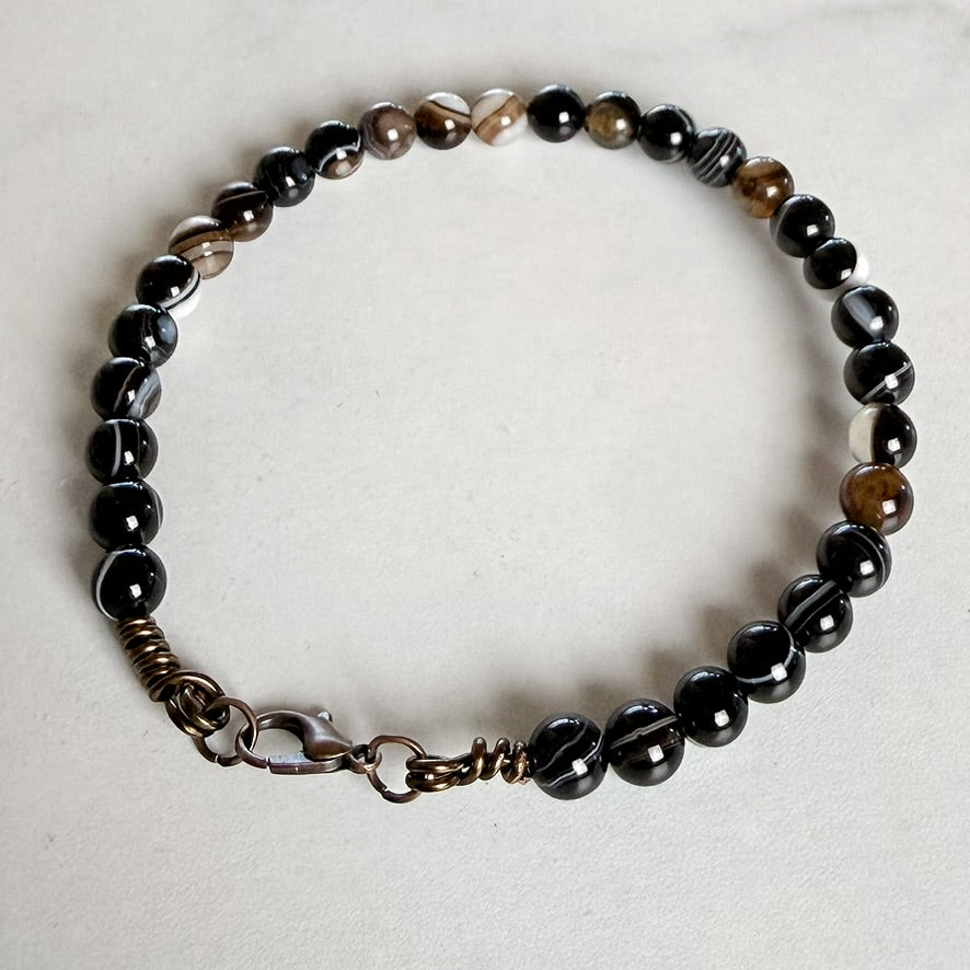 A beaded bracelet made of glossy black beads with some brown and white marbled accents, featuring a bronze-toned clasp on a light gray background.