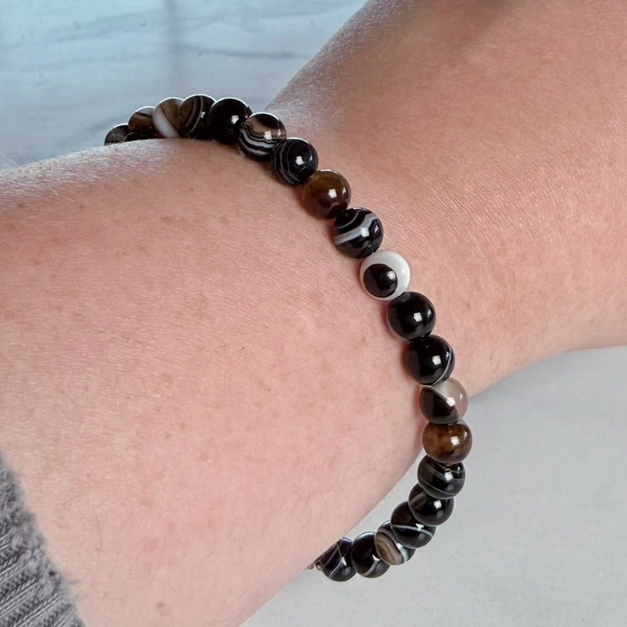 A beaded stretch bracelet worn on a person's wrist, featuring glossy round beads in black, dark brown, tiger-eye brown, and white marbled patterns.