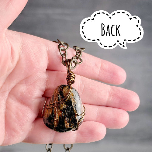 A hand holding a small pendant necklace with a rough stone pendant and antique bronze chain, with a speech bubble label reading 'BACK' on a gray background.
