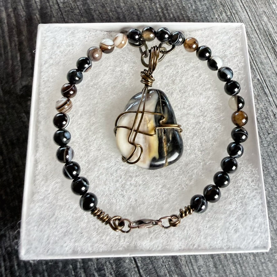 A beaded bracelet displayed on a white textured jewelry box. The bracelet features glossy black beads, several brown and cream marbled beads, and a large irregular polished stone pendant wrapped in bronze-toned metal wire.