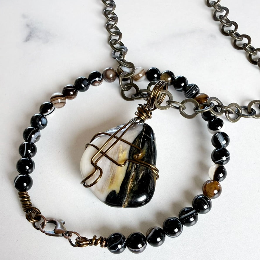 A beaded necklace with a central polished stone pendant wrapped in copper wire, displayed on a light marble surface. The necklace features black, white, and brown beads with a dark metal chain.