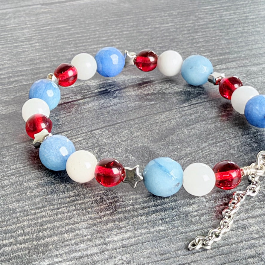 A beaded stretch bracelet featuring light blue, white, and red beads, accented with a silver star charm and a silver chain extender, displayed on a gray wood-textured surface.