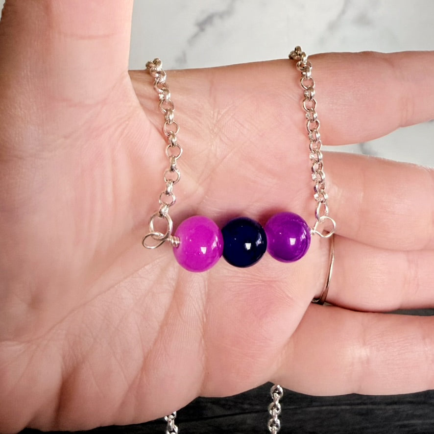 A close-up of a hand holding a delicate silver chain necklace with three glossy round beads in bright pink, deep navy blue, and purple.