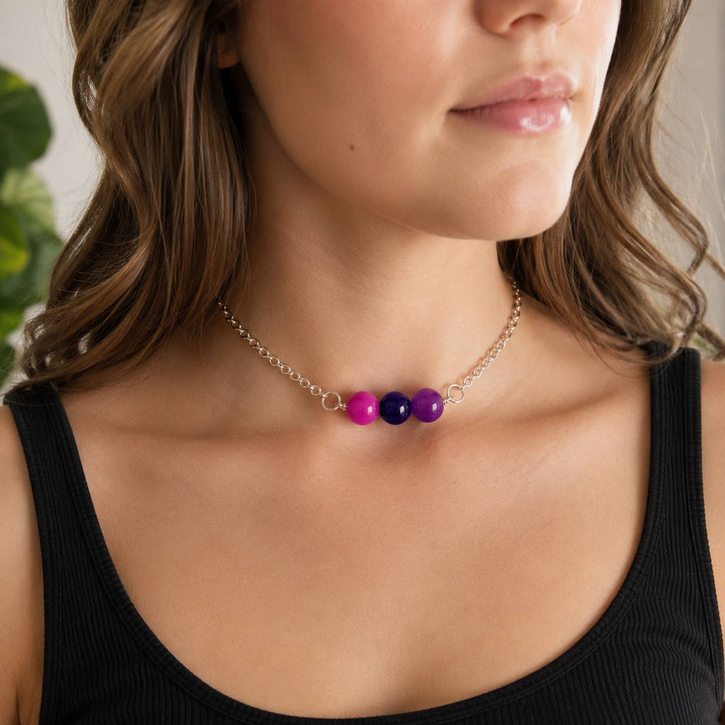 A close-up of a woman wearing a delicate gold chain necklace with three small glossy beads in pink, purple, and dark blue at the center, paired with a black ribbed tank top.