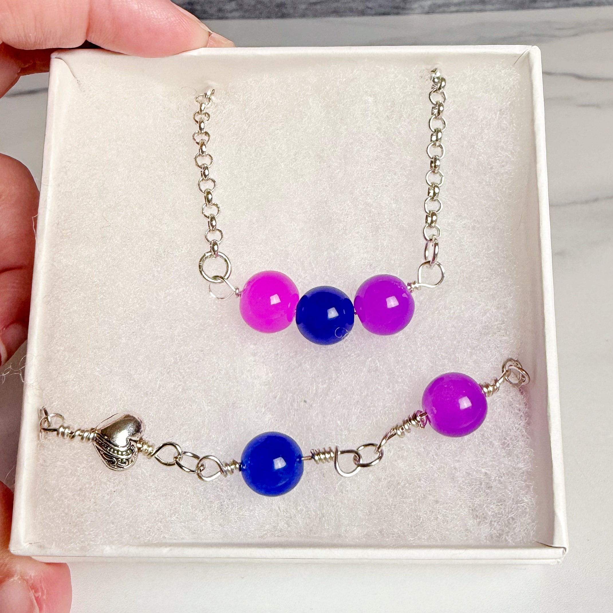 A jewelry set displayed inside a white gift box, featuring a short silver chain necklace with three beads (two pink, one blue) and a matching bracelet with a silver heart clasp, blue and pink beads, and silver links.