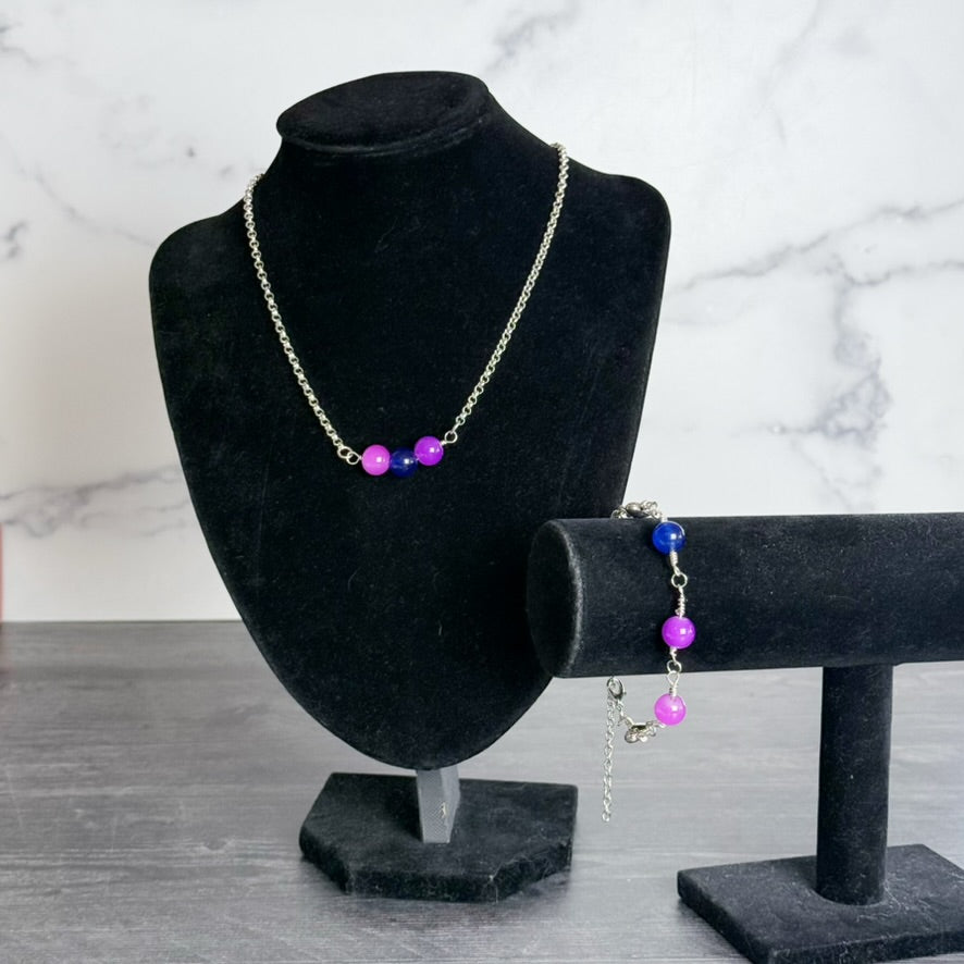 A silver-tone necklace and bracelet set with purple and blue beads, displayed on black velvet jewelry stands against a white marble-patterned background.