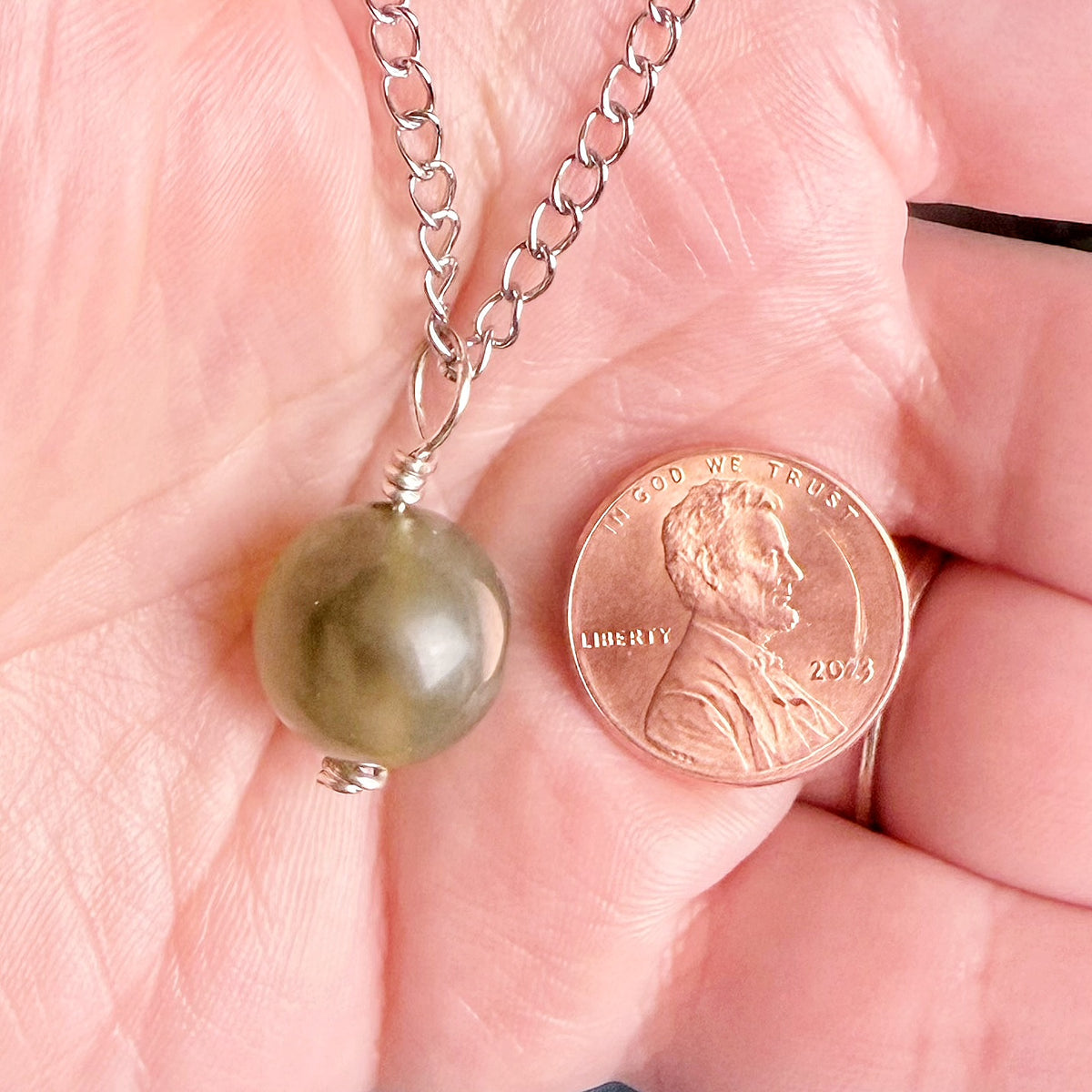 A silver chain necklace with a round olive-green bead pendant held in a person's hand next to a U.S. penny for scale.