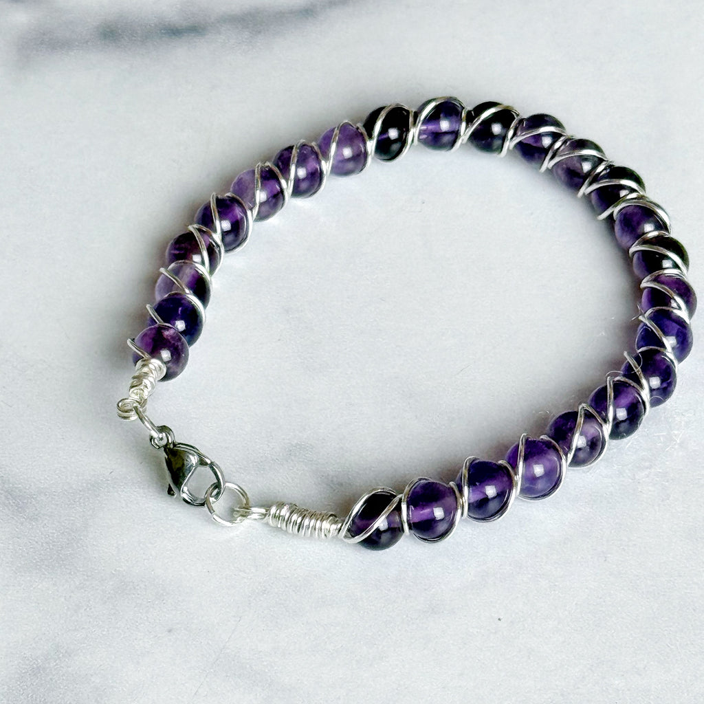 A beaded wire bracelet featuring polished purple amethyst gemstones wrapped in silver-toned metal, with a silver lobster clasp, displayed on a light marble surface.
