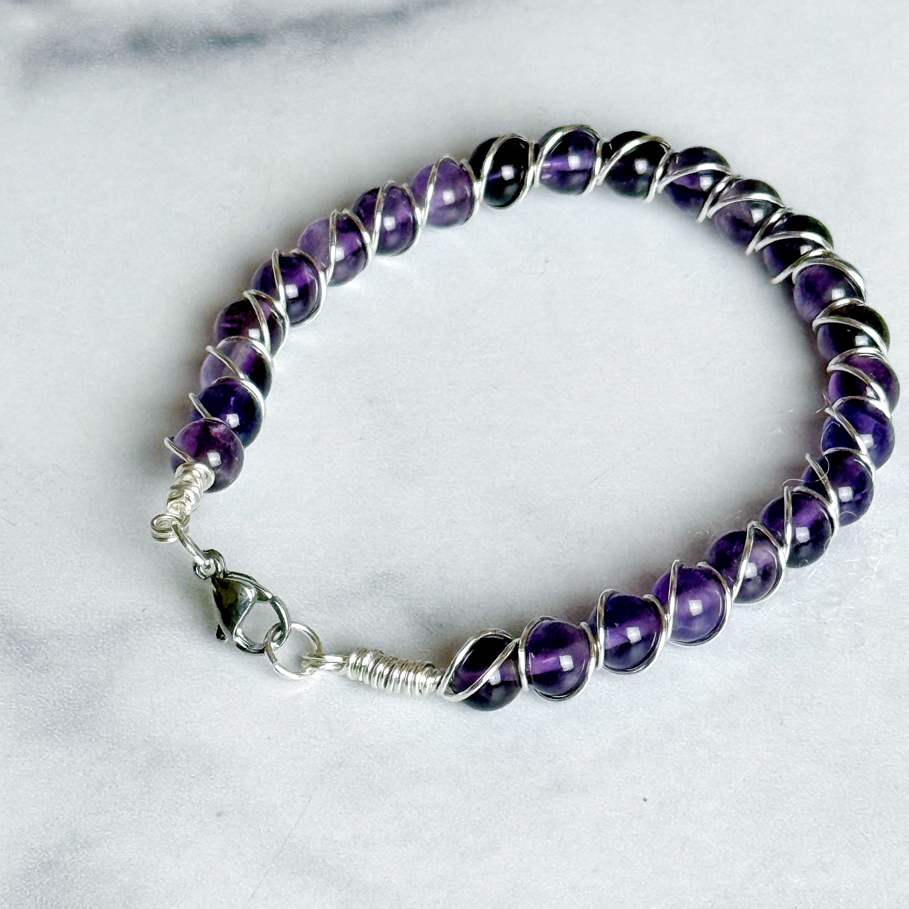 A beaded wire bracelet featuring polished purple amethyst gemstones wrapped in silver-toned metal, with a silver lobster clasp, displayed on a light marble surface.