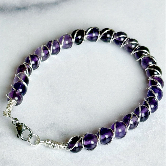 A beaded wire bracelet featuring polished purple amethyst gemstones wrapped in silver-toned metal, with a silver lobster clasp, displayed on a light marble surface.