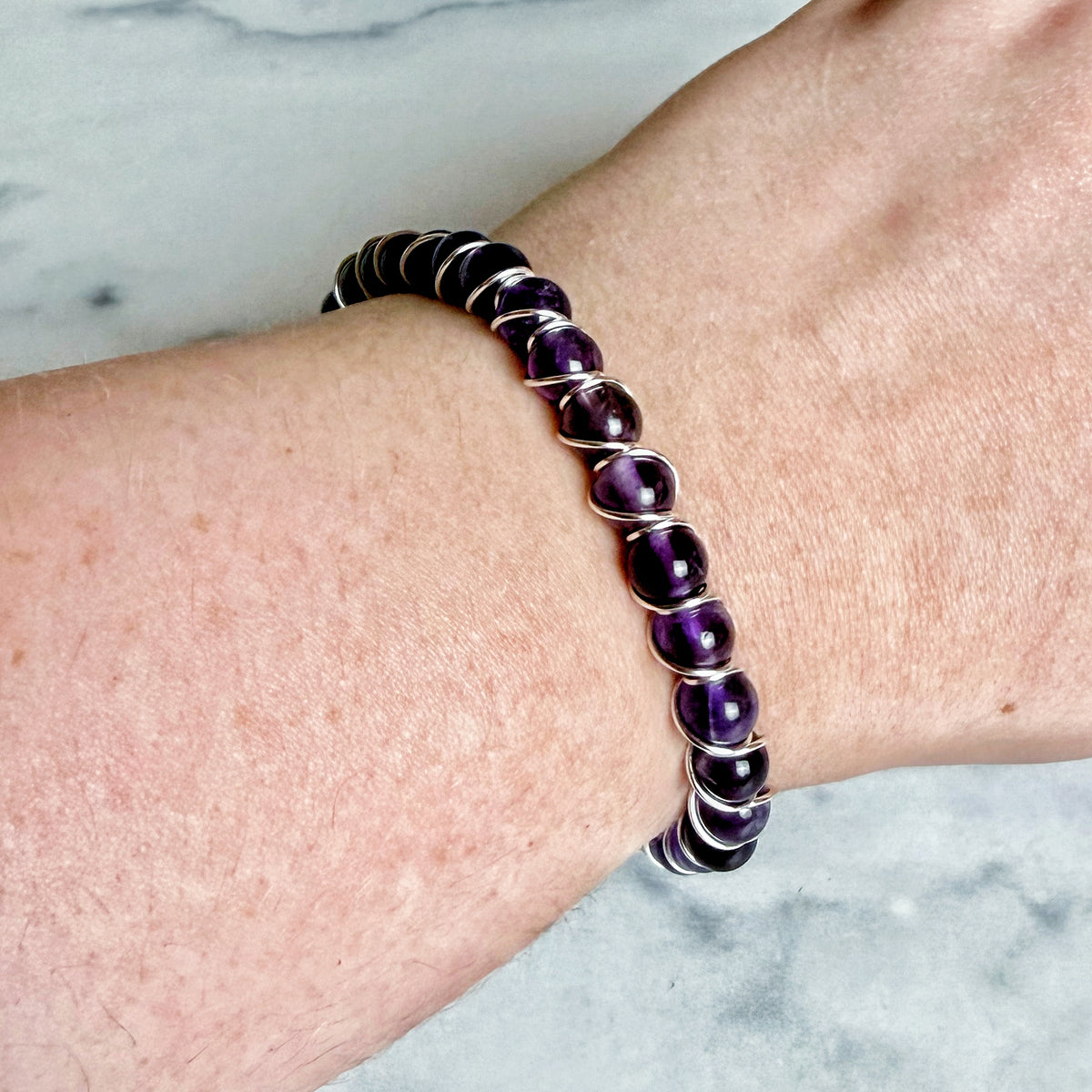 A close-up of a person's wrist wearing a beaded stretch bracelet made of polished purple oval beads separated by silver-toned metal rings, set against a light marble background.