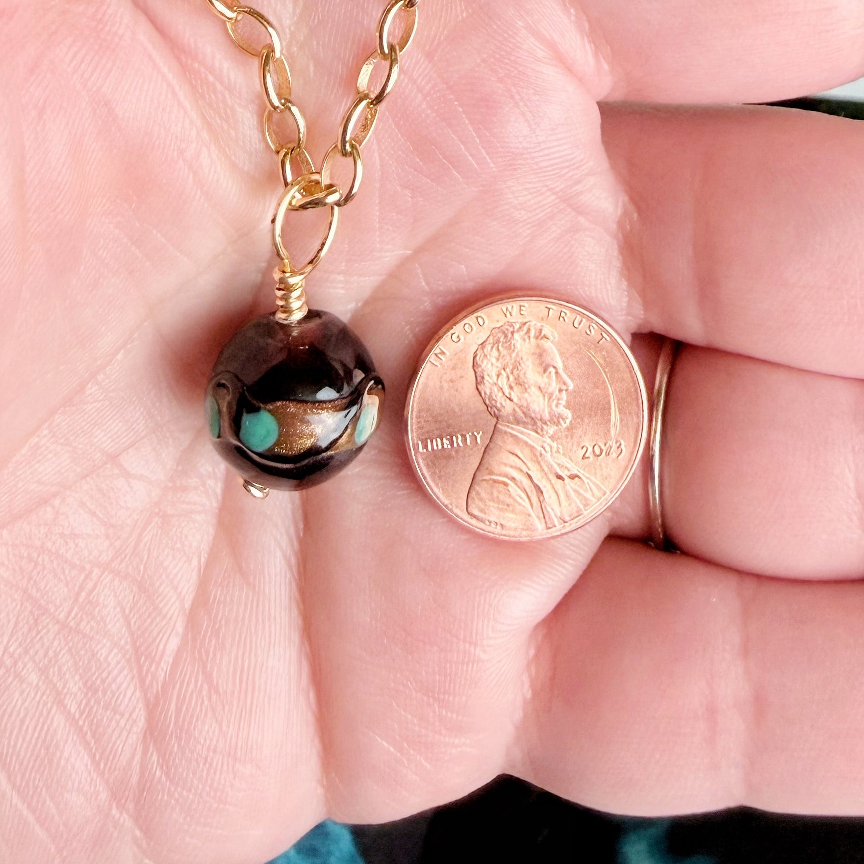 A close-up of a hand holding a gold chain necklace with a dark brown and turquoise glass bead pendant next to a US penny for scale.