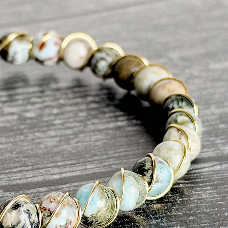 A close-up of a beaded bracelet made of round natural stone beads in white, gray, brown, and pale blue, wrapped in gold-toned metal, resting on a dark wood surface.