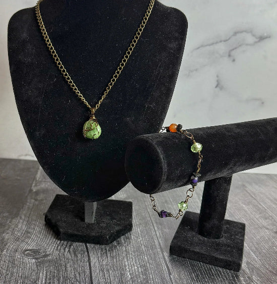 Necklace and bracelet with green and purple stones on black stands against a marble background