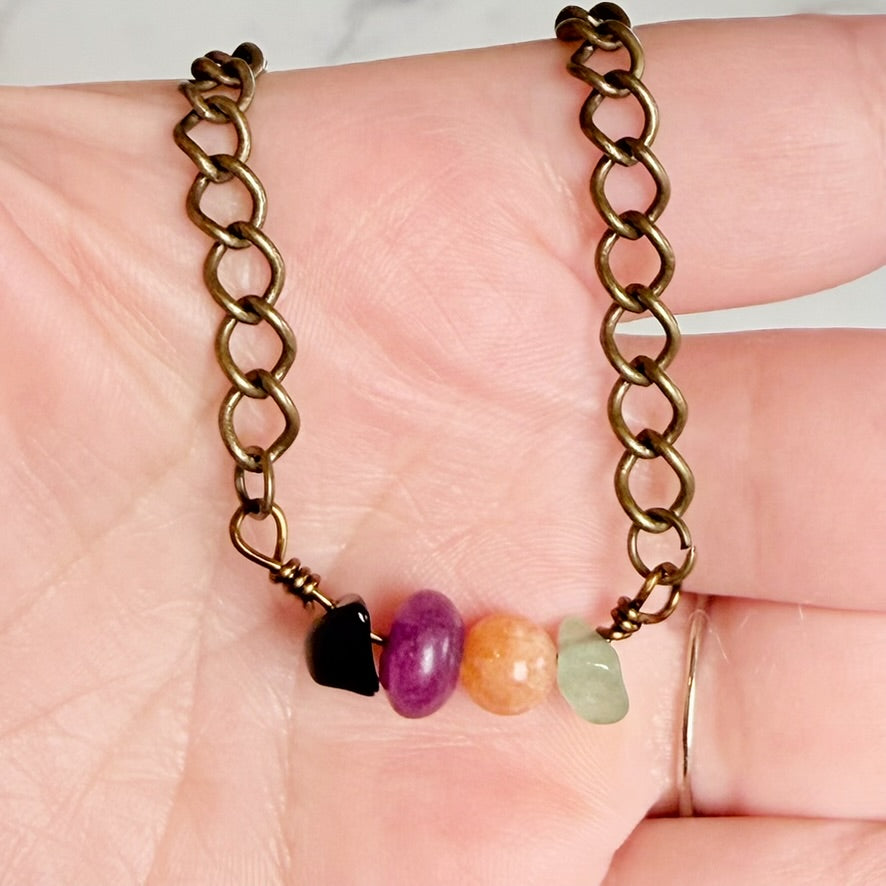 A close-up of a bronze chain necklace draped across a person's palm, featuring four small polished gemstone beads in black, purple, orange, and light green.
