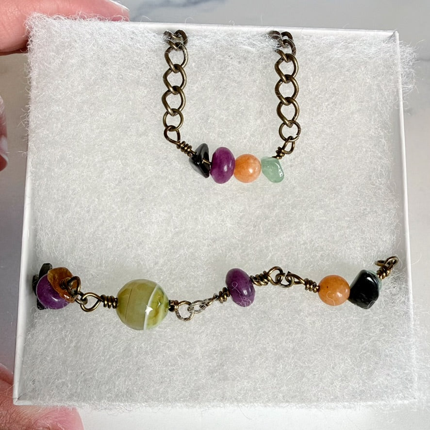 A jewelry set displayed on a white foam insert, featuring a short chain necklace and a longer beaded bracelet with antique bronze-toned metal and multicolored beads including purple, orange, green, and black.